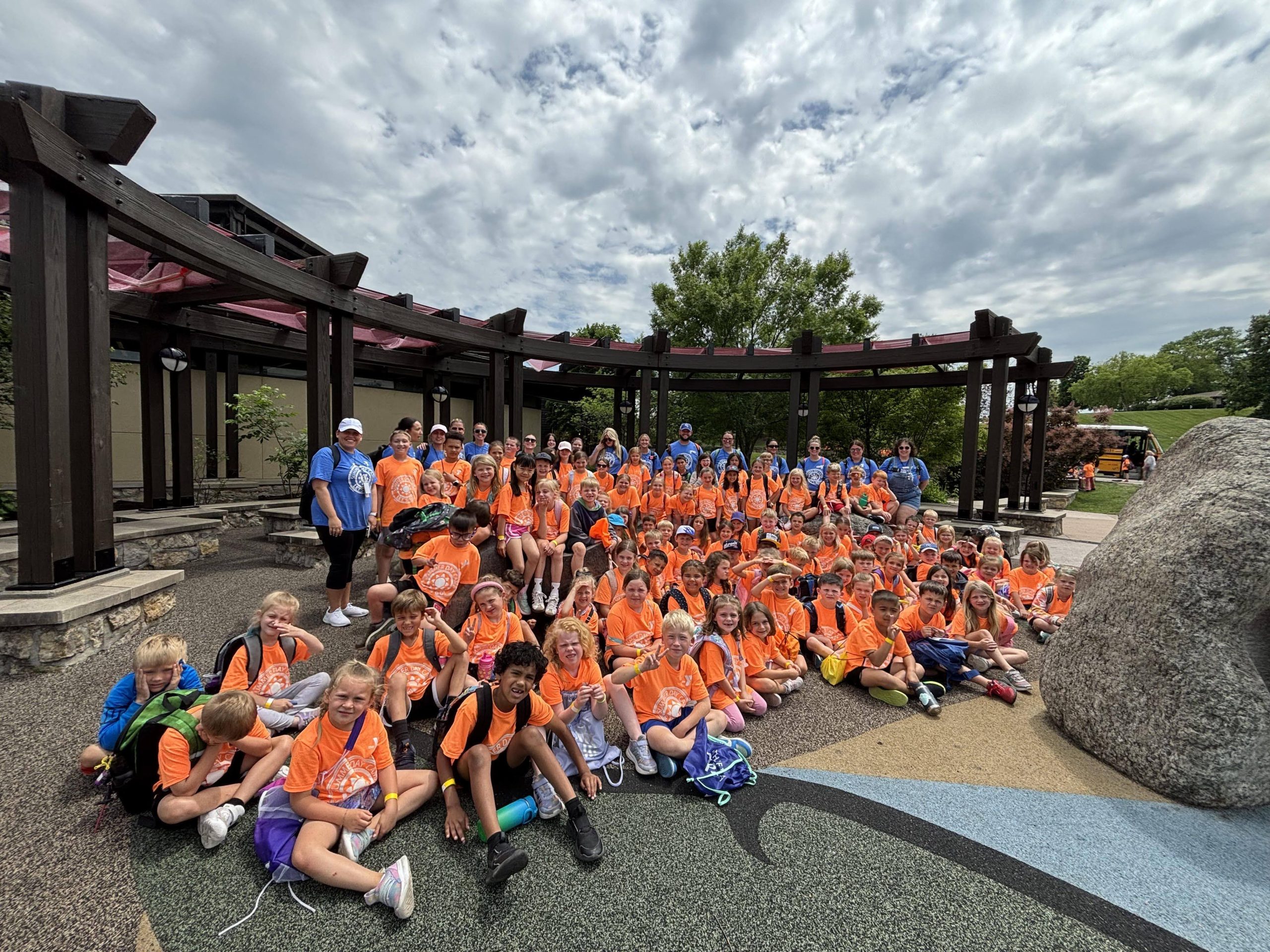 Large group of summer campers in orange shirts posing outdoors under a pavilion