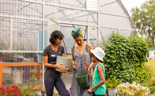 Woman staff member with a mother and child gardening outside a greenhouse holding a green watering pail