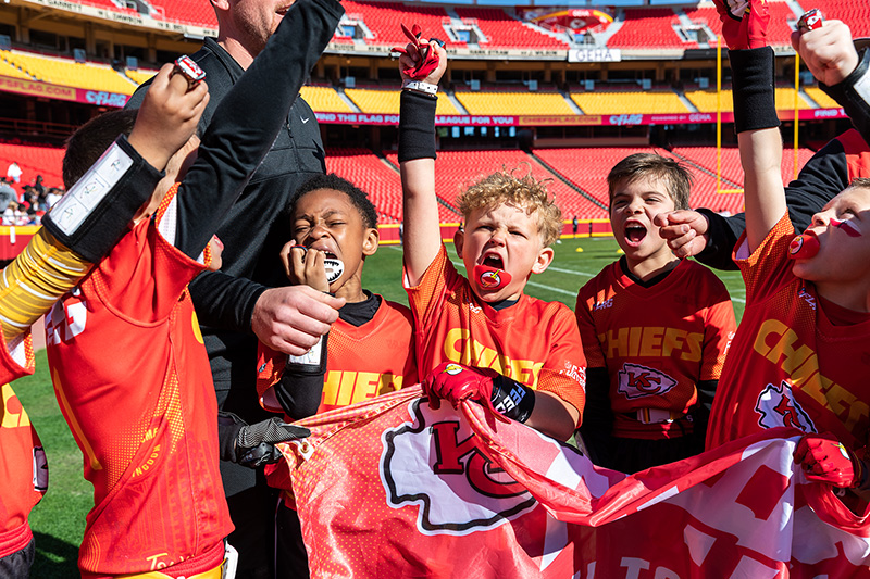 Youth flag football players in Chiefs jerseys cheering together on a stadium field