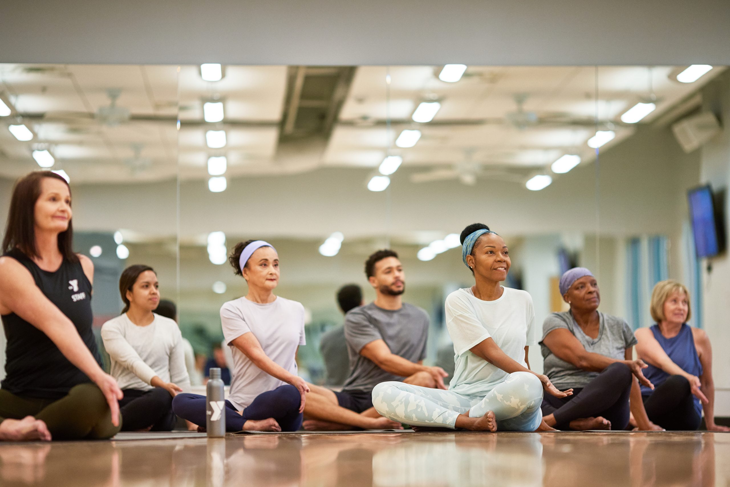 Diverse group of adults seated on the floor of a mirrored studio smiling during a group wellness class
